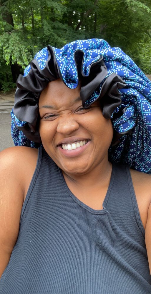 K in blue polka dot XL bonnet with black satin and large ruffles. She sewed this out of upcycled sheets. Second bonnet she made. She is in black tank and smiling. 