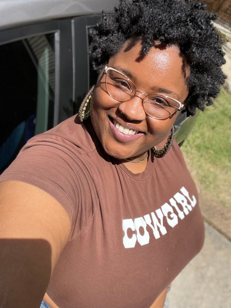 K in brown crop top that says COWGIRL in all caps. She is wearing brown and gold hoop earrings and glasses. She is smiling and standing next to her car. 