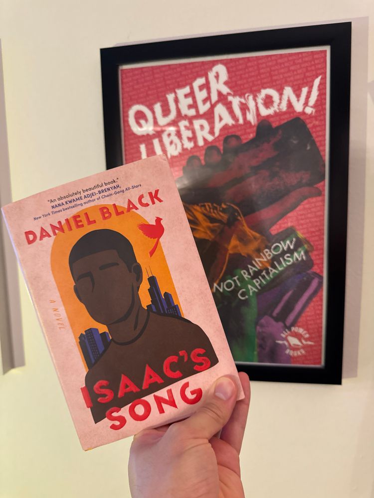 My hand holding Daniel Black’s latest novel “Isaac’s Song” up against the backdrop of a white wall and poster from All Power Books that reads “QUEER LIBERATION” in an electrifying larger font and “NOT RAINBOW CAPITALISM” in a smaller, somewhat glitchy font. 