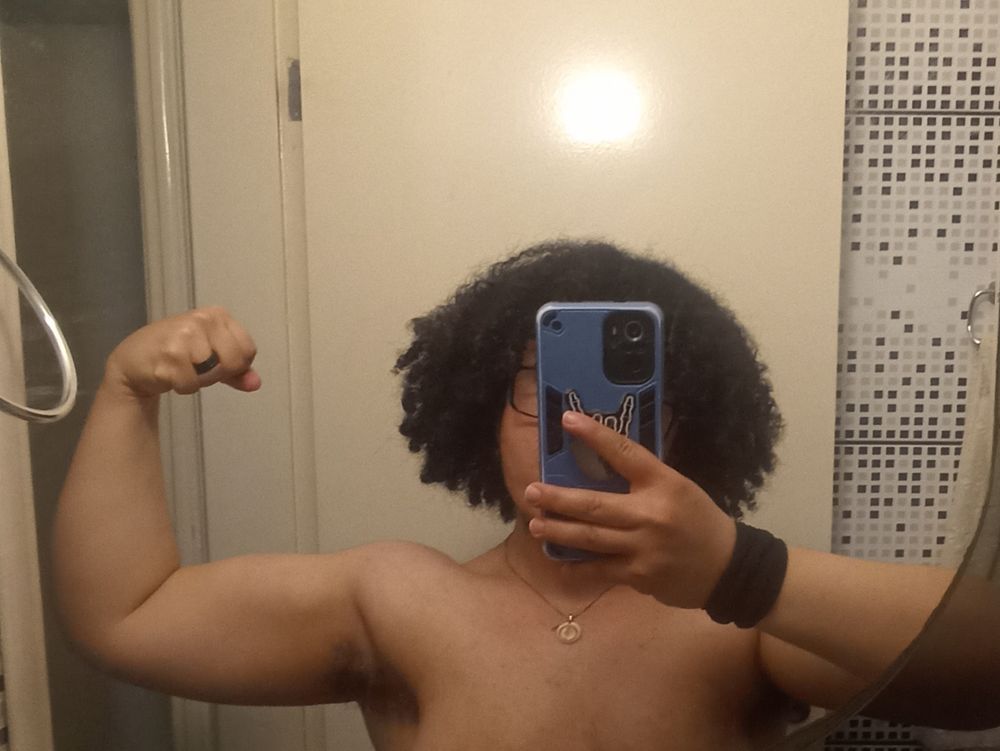 Foto minha posando os biceps. Estou sem camiseta, na frente do espelho. Cabelo escuro cacheado e volumoso acima do ombro, pele clara. Dá pra ver um pouco de volume no biceps e ombro. 