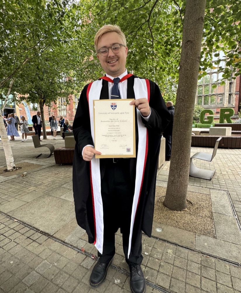 A picture of Daniel wearing a black suit, and black robes with a white and red sash. He has blonde hair, round glasses, and is holding up his degree certificate with a big smile. 