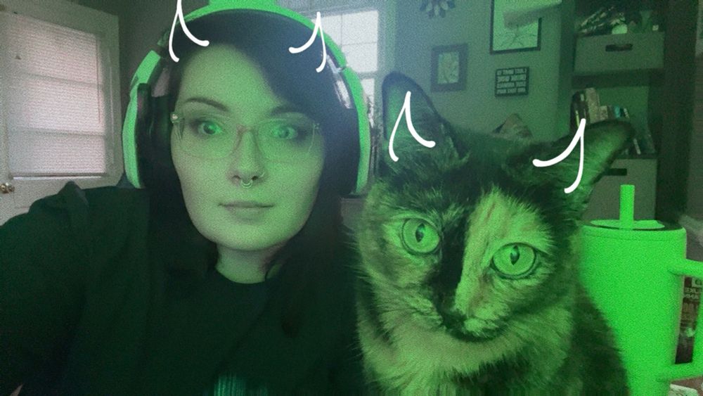 a selfie of me and my cat with the silly devil horns Snapchat filter. It’s green bc of my RGB office light 
