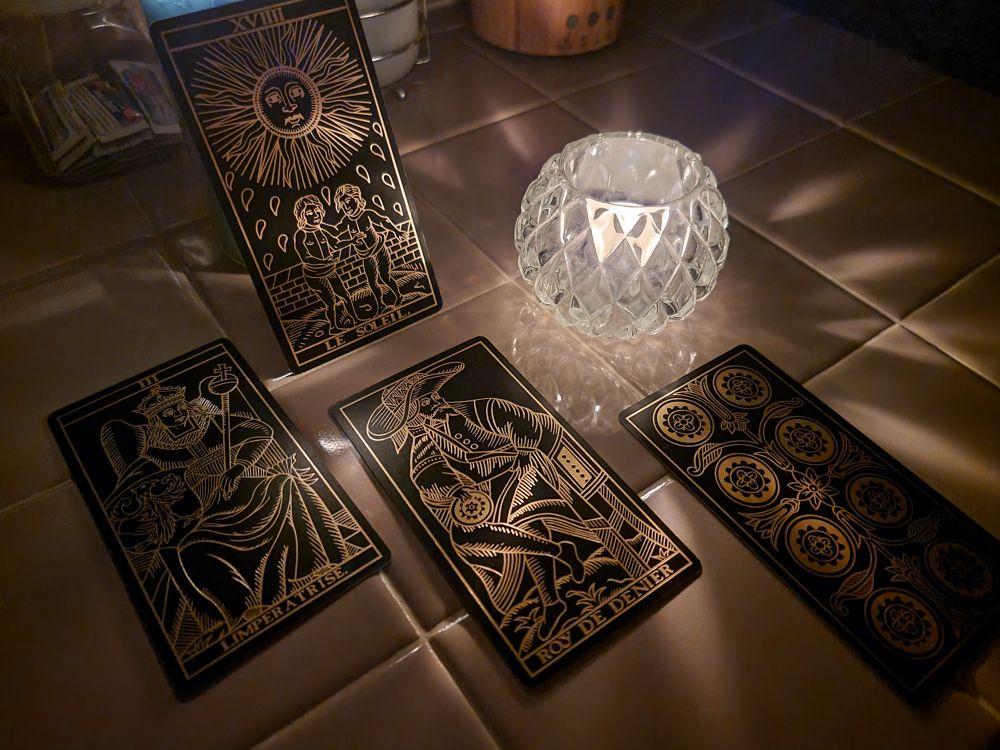 Four , black background with metallic gold embossed images from a Tarot de Marseille style deck, reflecting candlelight in a darkened room. 
