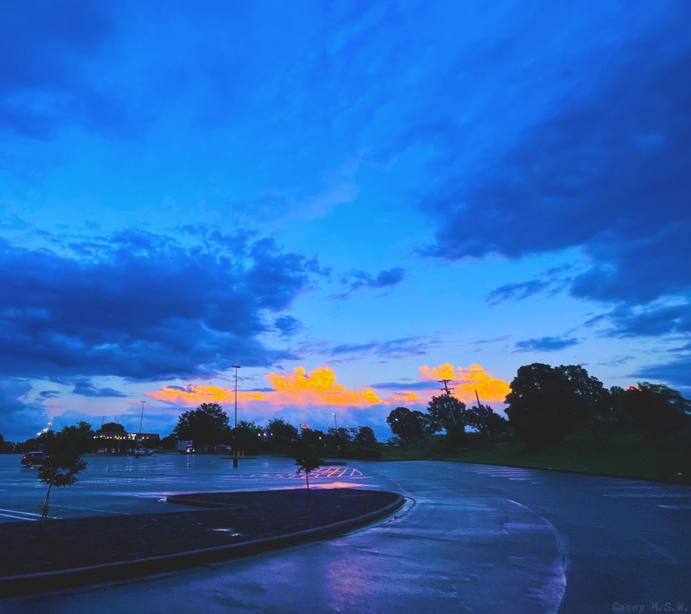 In a parking lot during the evening, a cellular tower, streetlights, utility poles, vehicles, and various establishments with the lights still on can be seen from afar, with trees of varied sizes and in different areas.

There's a reflection of the evening light from when it was raining on the pavement within the empty partaking lot itself.

The background also has orange and blue tones with stratocumulus and stratus clouds.