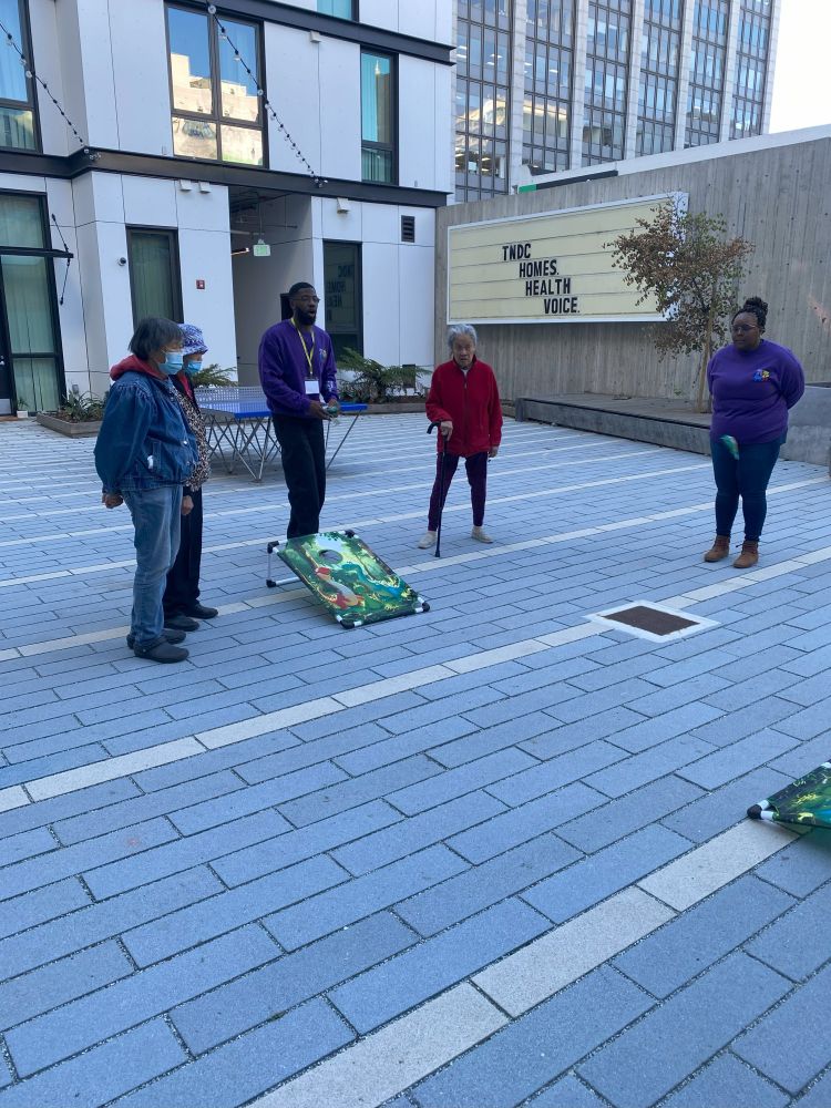 In a courtyard, two volunteers demonstrate how to play a bean bag toss game while three seniors watch. 