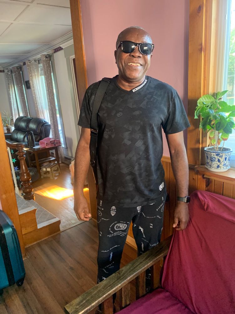 My dad, a Black man smiling, wearing black sunglasses black shirt and some kind of black and white patterned pants