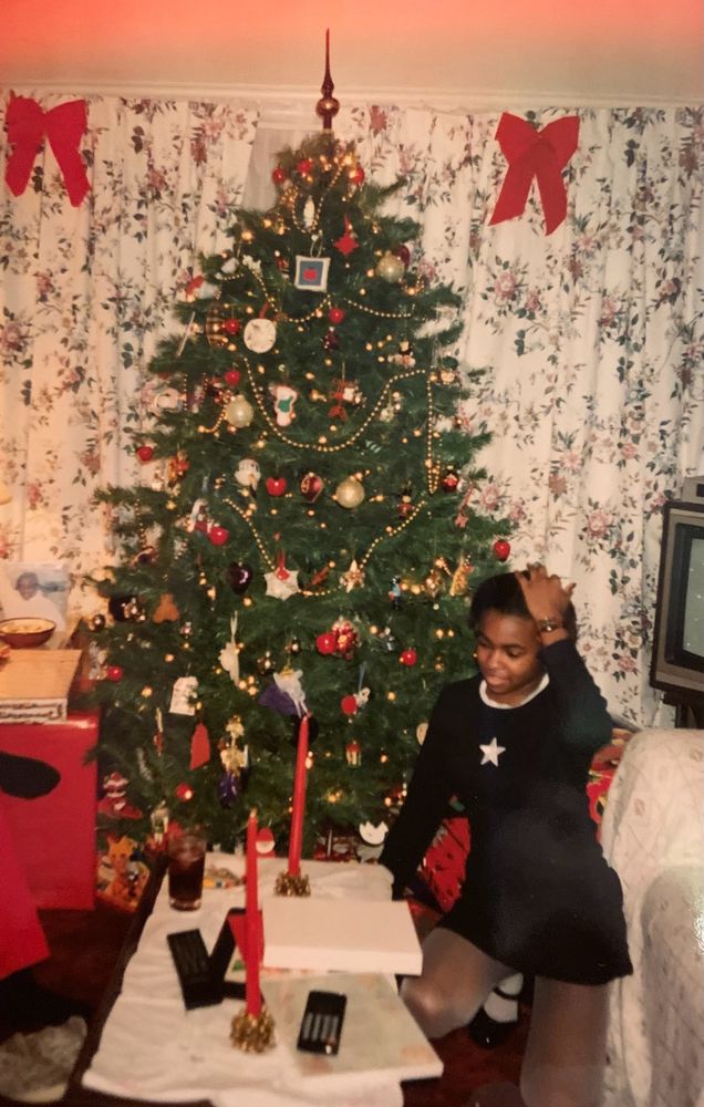 Black girl in living room with Christmas tree circa 1994 or 95 kneeling with hand on head wearing black mini dress with white star on chest and white trim around neck white panty hose and black patent leather Mary Jane shoes
