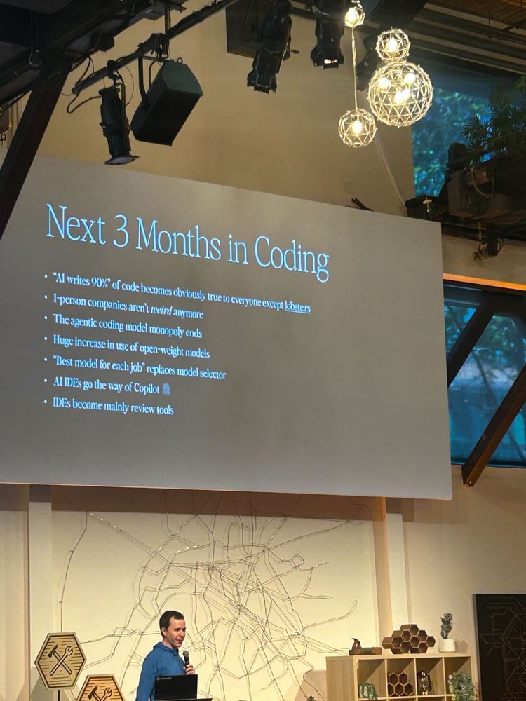Guy presenting at a tech conference; the title is 'Next 3 Months in Coding' and the top bullet point is '"AI writes 90%" of code becomes obviously true to everyone except lobste.rs' and yeah the weird quotes are in the original.