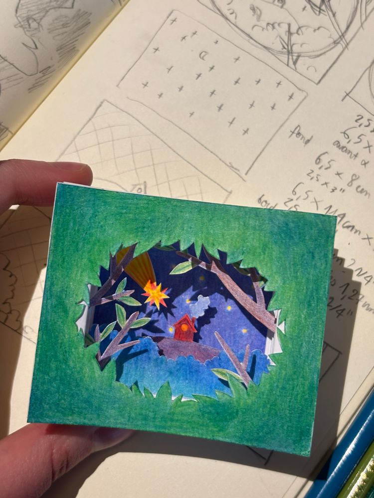 A picture of a small tunnel book: a scene representing a small house on a hill at night with a shooting star, seen through leaves and branches. The tunnel book is made out of different paper panels cut and glued together so as to simulate perspective. These are coloured with coloured pencils.