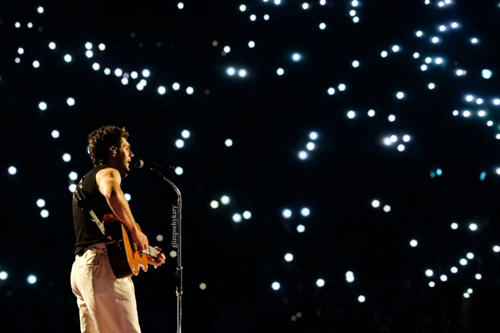 Niall Horan holding his guitare standing in front of his mic stand. The background is full of flashlights / torches from fans. His gaze is on the crowd. He is wearing white trousers and a black vest, standing sideways. 