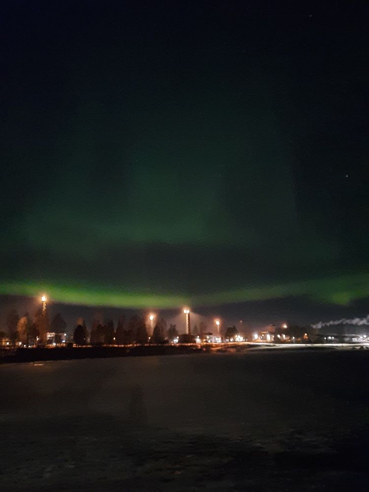 Vihreitä revontulia järven päällä. Vastarannalla näkyy kaupungin valoja.