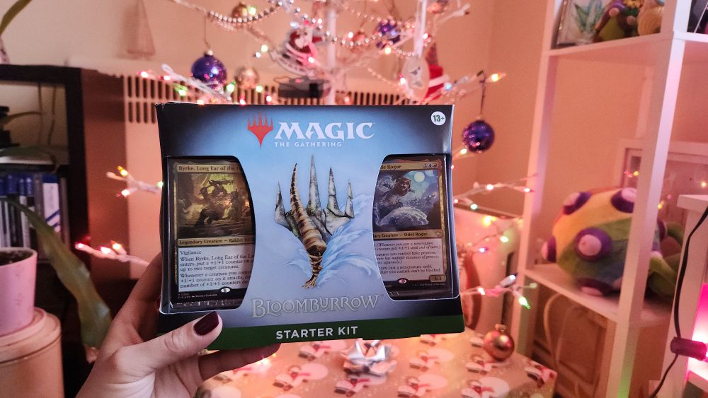 My hand holding up a Magic: the Gathering Bloomburrow starter set in front of our lit Christmas tree