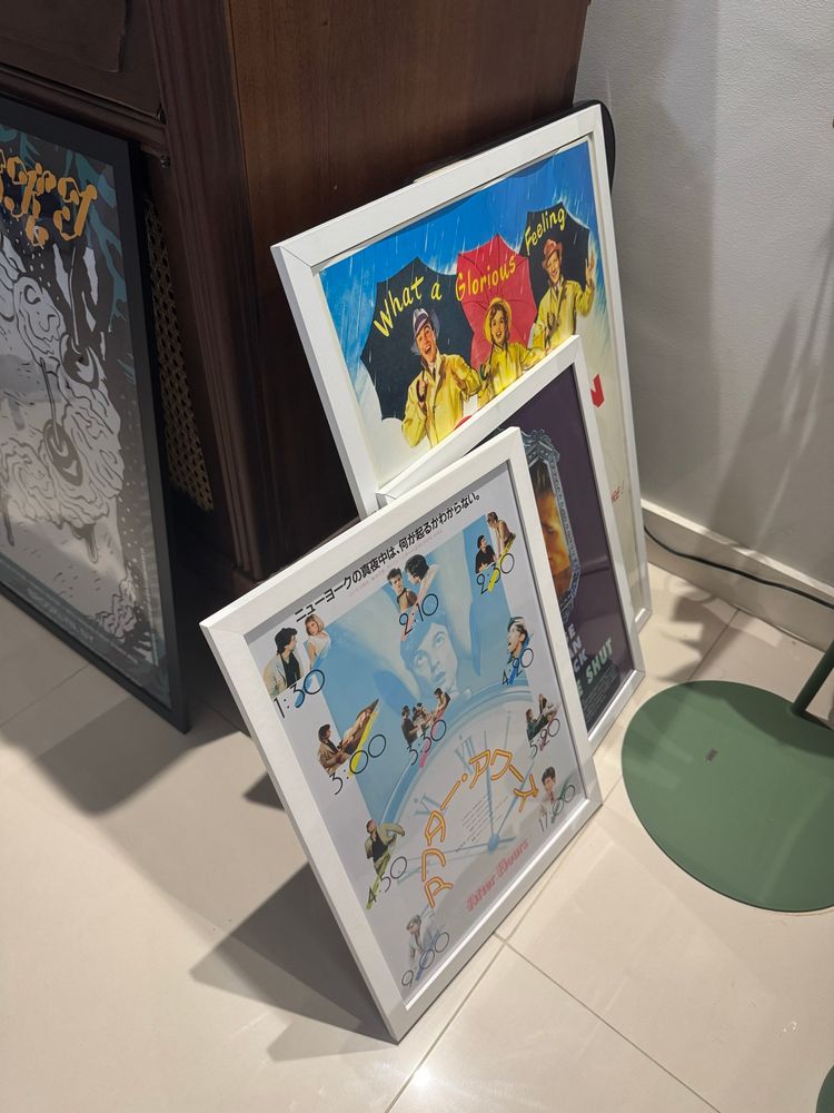 a foto mostra um móvel de madeira. apoiado nele está um pôster grande da mitski com moldura preta, um pôster grande de cantando na chuva com moldura branca, e dois pôsteres médios em moldura branca (um de after hours e um de eyes wide shut)