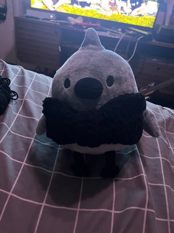 small gray crow-like plushie known as a doodle plushie is wearing a black crocheted bow tie sitting on grey and white grid pattern bed sheets