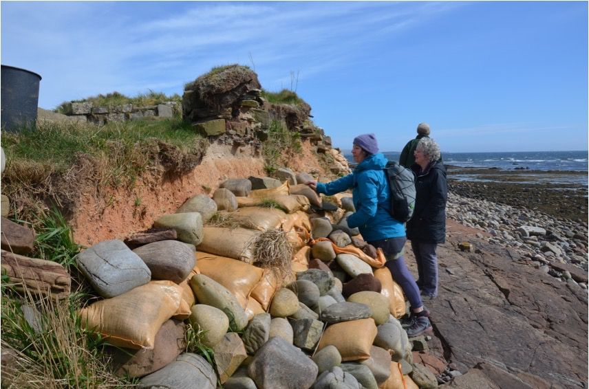Three people on a rocky beach examine a low eroding bluff that has been temporarily shored up with sandbags  The site is a Medieval cemetery on Mainland, Orkney. Photo: Adam Markham