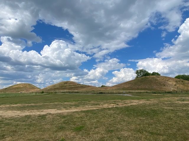 Three grassy Iron Age burial mounds at Gamla Uppsala against a cloudy blue sky. Grass in the foreground.