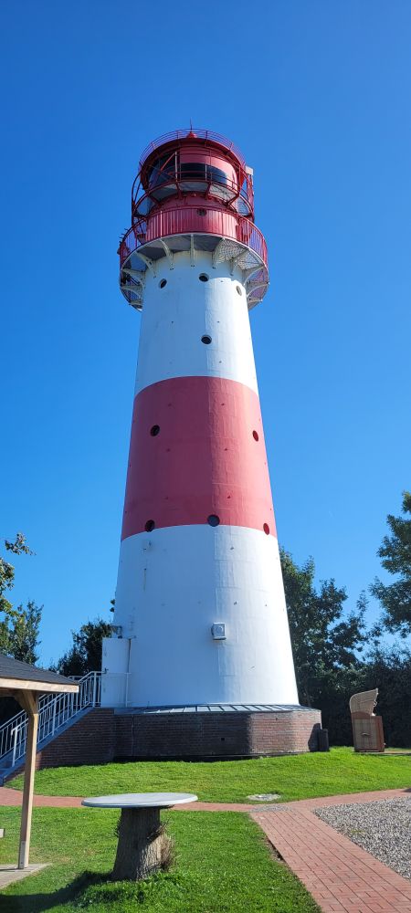 Foto eines rot/weiß  gestreiften Leuchtturms von vorne.  Der Himmel dahinter ist strahlend blau und wolkenlos. Man sieht Wege vor dem Leuchtturm (rote Ziegel) und links im Bild einen Tisch und die Ecke einer überdachten Sitzgelegenheit.
