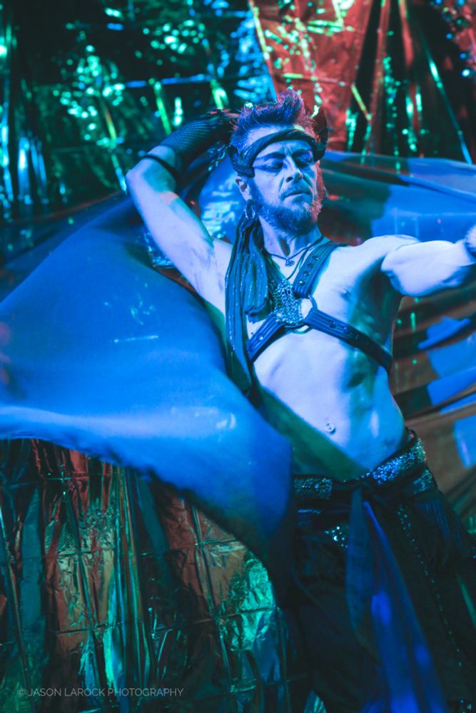 A masculine belly-dancing performer with a large, sheer piece of fabric fluttering behind them. 