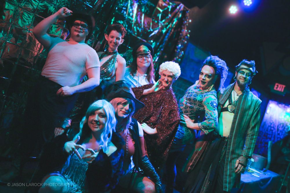 A group of drag performers are gathered together on stage, smiling towards the camera. Each one is uniquely dressed for their stage persona. 