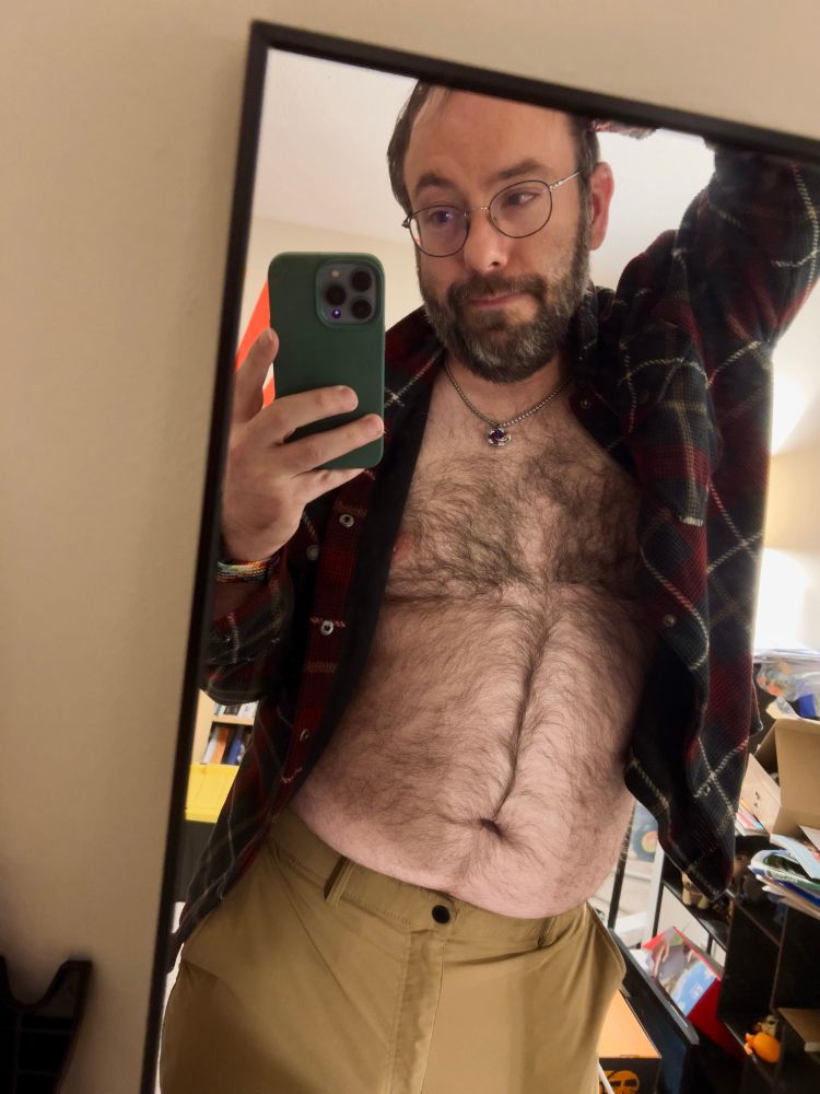 Selfie of me in the corner of a mirror. I am wearing. a buttoned up plaid shirt that’s open to expose my chest and brown pants. My arm is raised above my head.