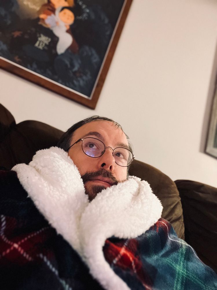 Selfie of me shrouded in a plaid blanket with fuzzy white liner. My head is peaking up just barely. 