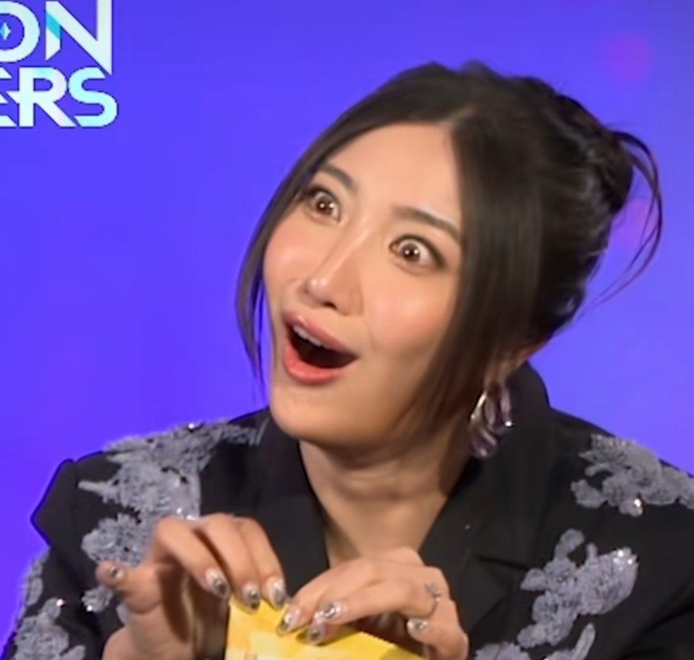 Ejae in front of a purple background with a surprised and annoyed look on her face. Her fingers are delicately holding a yellow bag just out of frame.