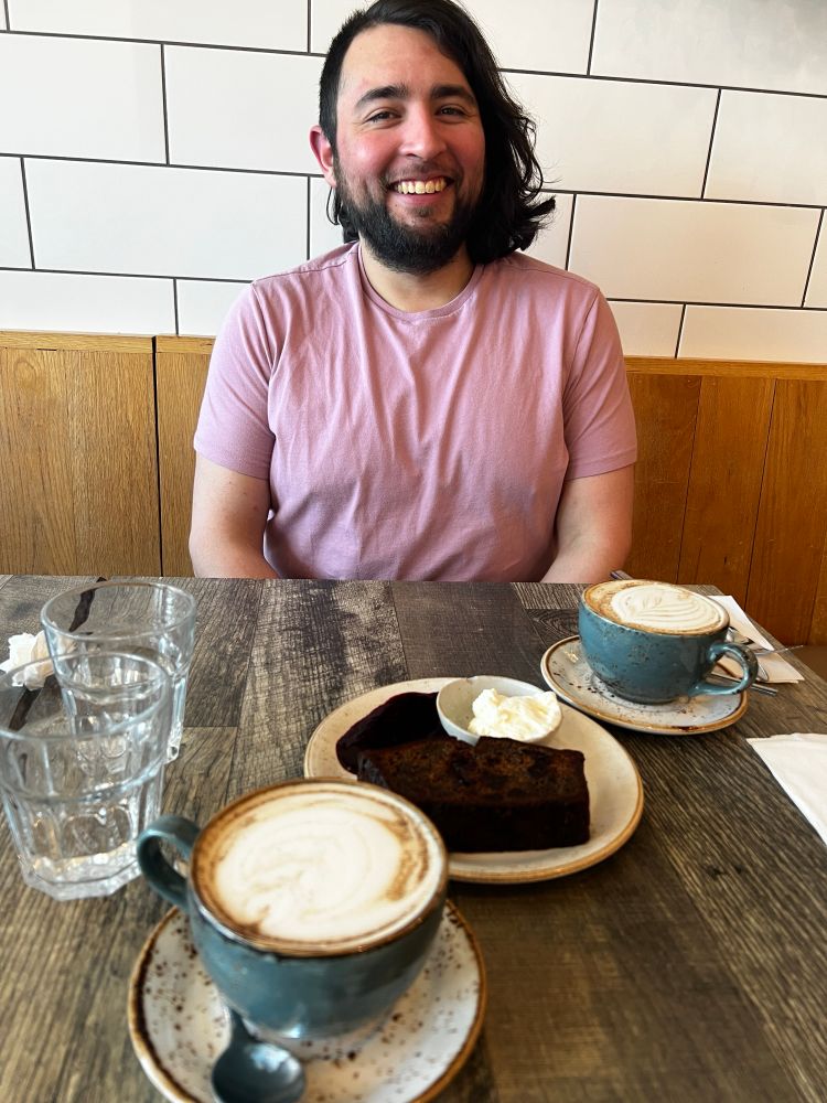 My boyfriend wearing a light pink shirt sitting on the other side of a table. There are two full cups of chai and a slice of banana bread between us.