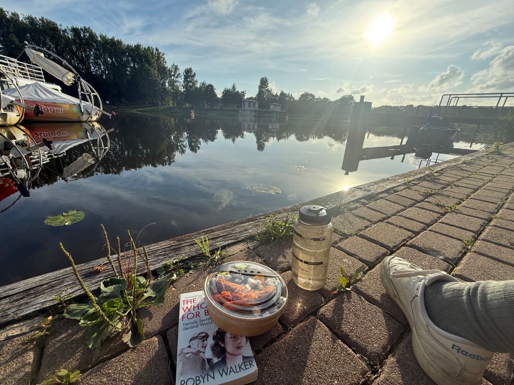 Sonnenuntergang am Kanalufer, bereit steht eine Dose mit Salat, eine Flasche Wasser und ein Buch (The women who spied for britain). Es ragt ein Fuß ins Bild. 