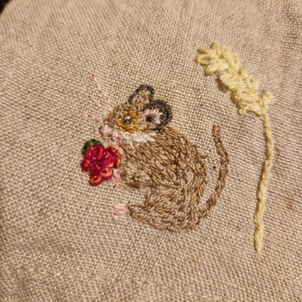 Embroidered brown mouse, sat on hind legs, eating a raspberry with a wheat strand nearby, all sewn onto brown linen