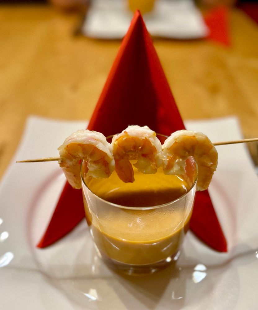 A skewer with three shrimp rests on a glass of gazpacho, placed on a white plate with a folded red napkin in the background.