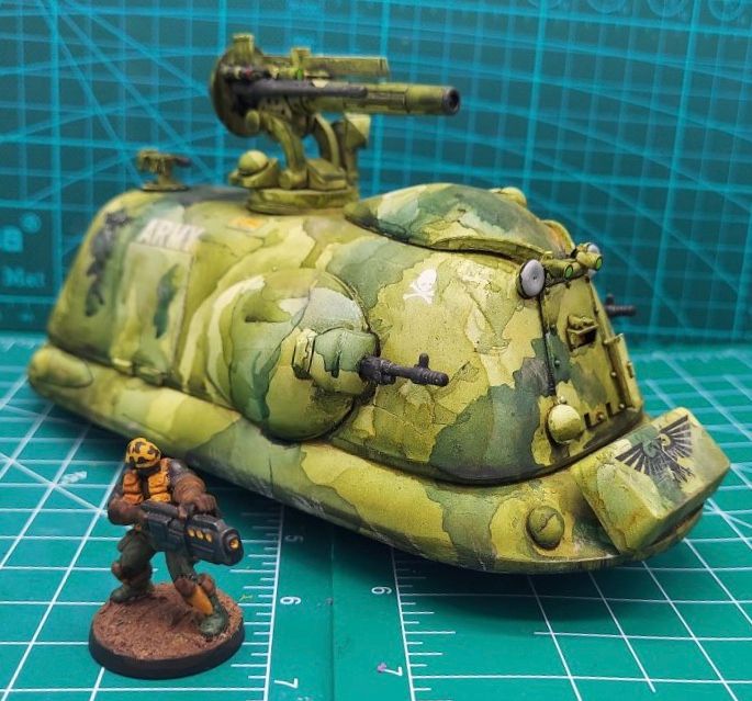 A finished science fiction hover tank made entirely out of recycled plastic bits and bobs, painted in various shades of green camouflage and alongside a 28mm miniature soldier. 