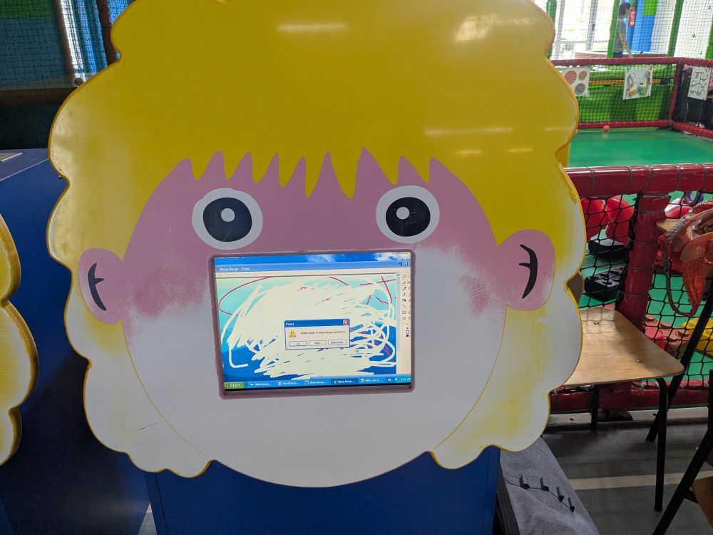 A kiosk computer screen in a (child friendly/scary) face shaped frame displaying MS Paint on Windows XP