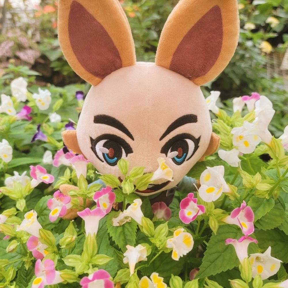 An image of the Gardener Skye plush smelling pink and white flowers in a garden.
