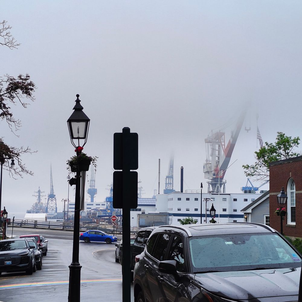 An image of cars driving down the road in downtown Bath, Maine. In the background are large cranes from the nearby shipyard partially obscured by fog.