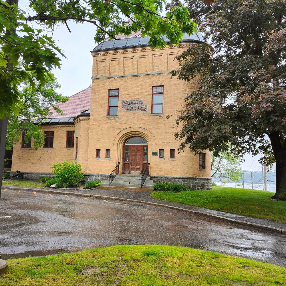 The front of the historical portion of the Patton Free Library. It is a large, tan brick building with a red roof and a dark wooden double door.