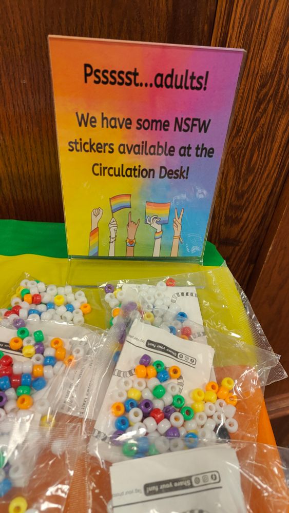 A table with a plastic rainbow tablecloth. There are some clear plastic bags that contain kits to Make rainbow beaded bracelets. There is a sign in the corner of the table that says, "Pssssst... Adults! We have some NSFW stickers available at the circulation desk!" The sign has a rainbow gradient background.