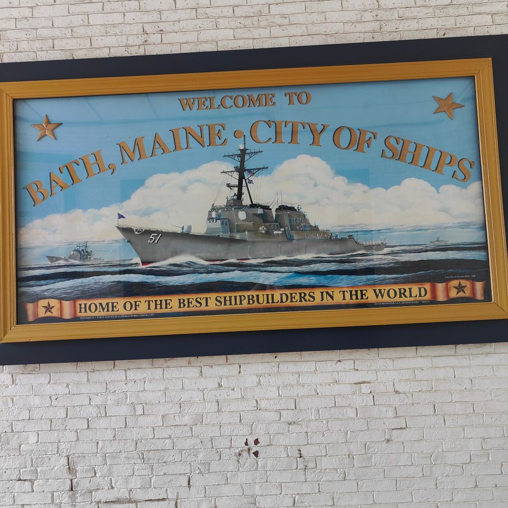 A framed sign against a white brick wall. The sign reads, "Welcome to Bath, Maine - city of ships - home to the best shipbuilders in the world." A destroyer class ship is in the center of the sign.