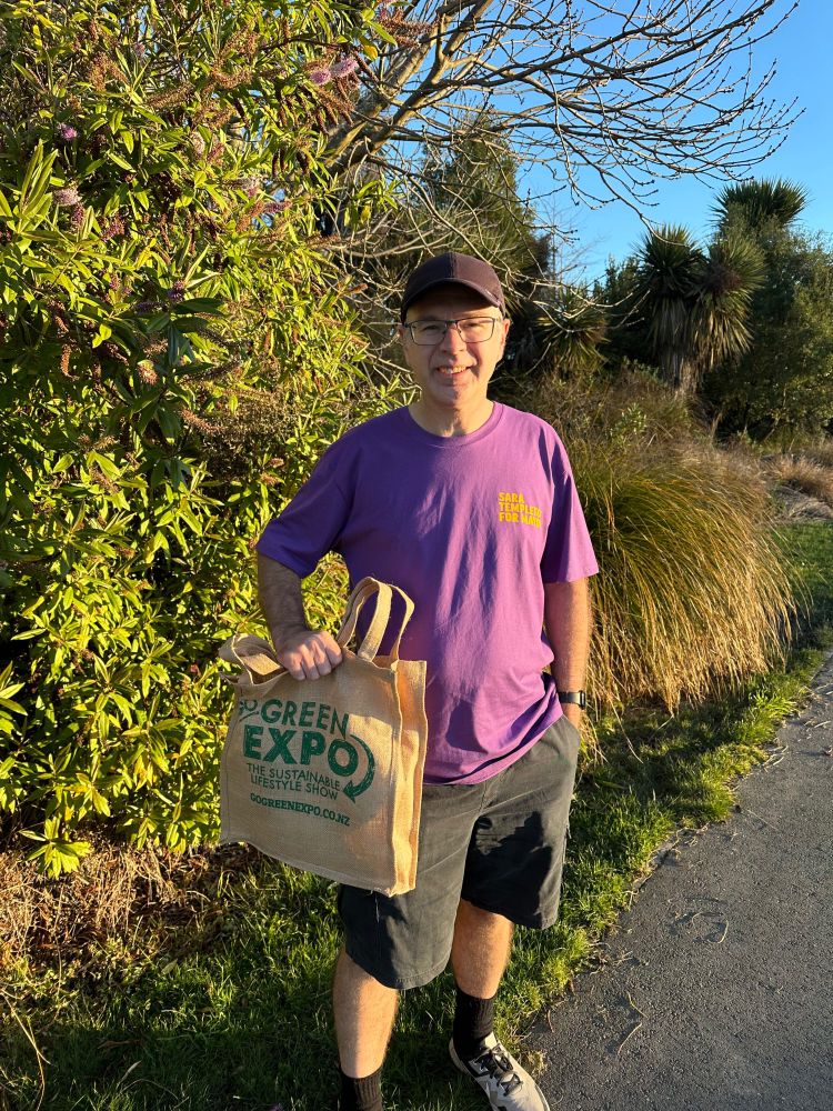 Allan stands on the edge of a path in his purple shirt in front of a bush/tree. He hold his prize bag (with the words Go Green Expo written on it) in his right hand and smiles for the camera. The sky is blue in the background. 