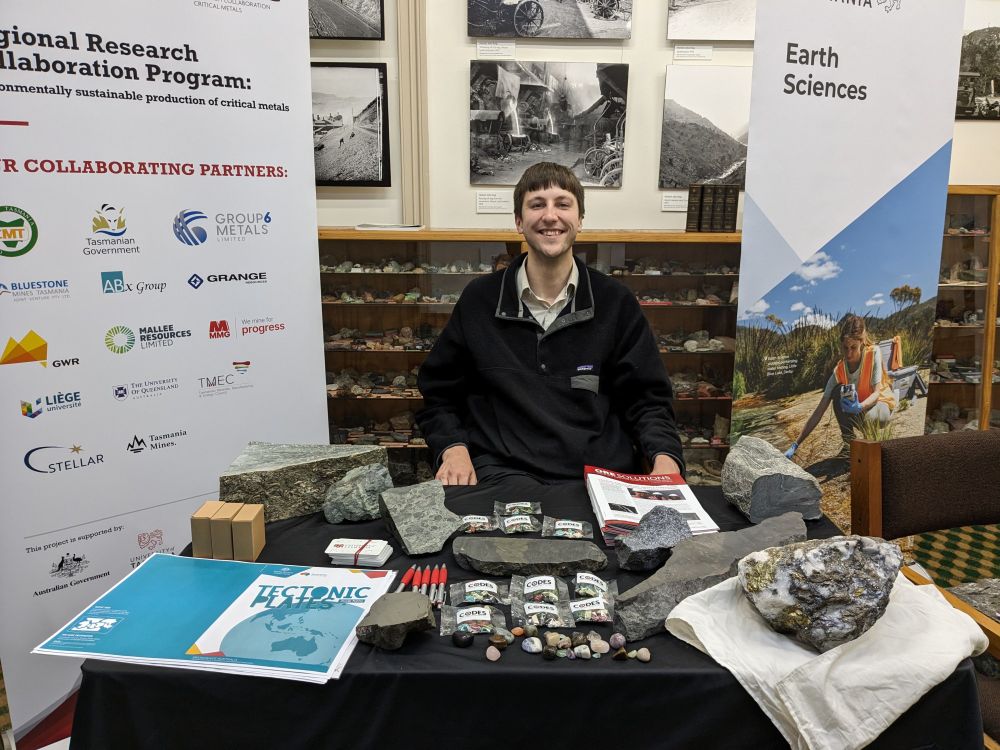 I am wearing a black jumper, sitting in front of a mineral collection and behind a table with various rock specimens from the University of Tasmania collection. Banners from University of Tasmania projects are visible on either side.