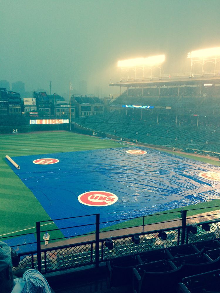 A baseball stadium with a blue tarpaulin spread over the infield. On each corner of the tarp is the Cubs logo. The field light are obscured by rain.