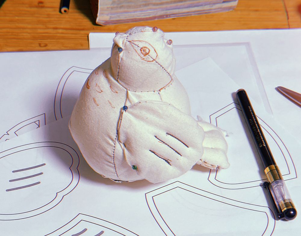 生成りの布で仮で作られた雷鳥のぬいぐるみ
スレッドひとつ前のポストの画像の型紙の上にぬいぐるみが頭を画面左に向けておいてあります。
仮なので模様などはペンで書いてあり、頭や羽の付け根など蟲ピンを使用して形を調整した跡があります。
画面の右側に置いてあるのはチャコペンです。