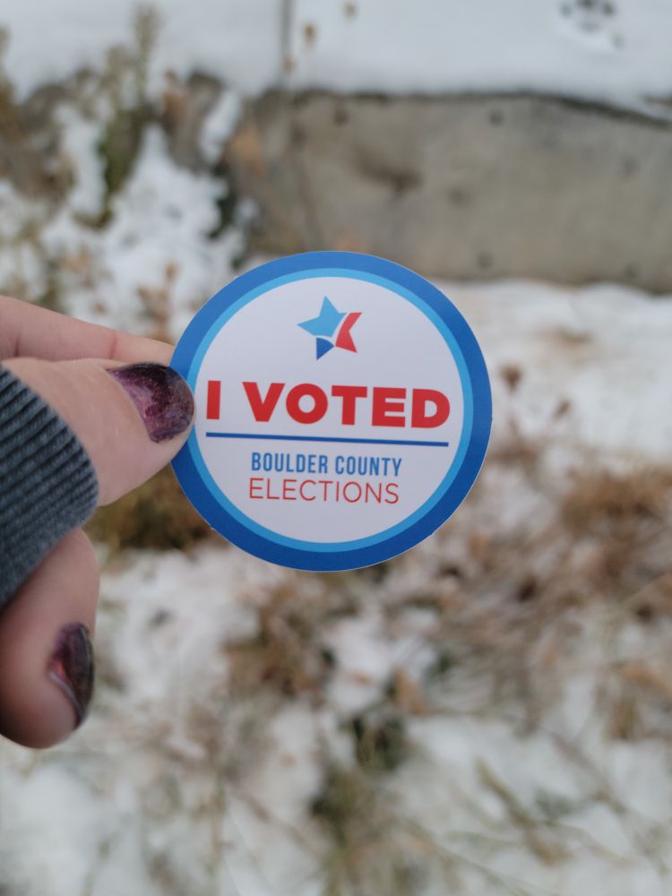 Round white sticker with a blue border. Red text saying I Voted with blue line underneath. Under the line blue and red text saying Boulder County (blue) Elections (red)