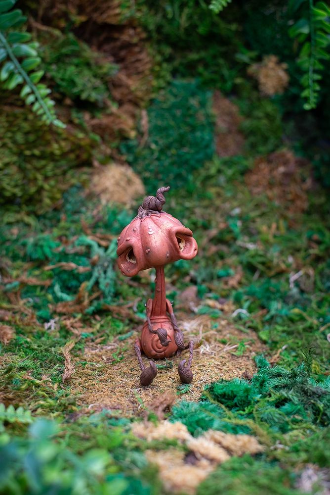 FRONT VIEW: Handmade polymer clay jack o lantern figurine sculpted in Mishes art style of oversized head with huge, sad eyes, thin neck & limbs, and a plump belly. Sitting on a moss background.