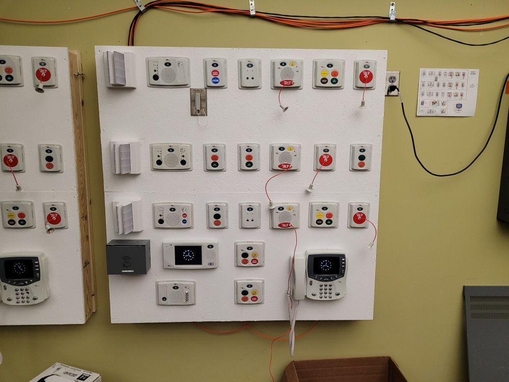 A completed white wooden test board with different nurse call modules attached and strung together in a mock environment for nurse call instalation training.