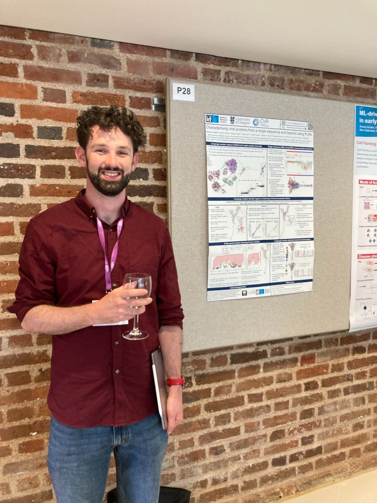 A man stands in front of a brick wall and a bulletin board displaying scientific posters and is holding a glass of wine. 