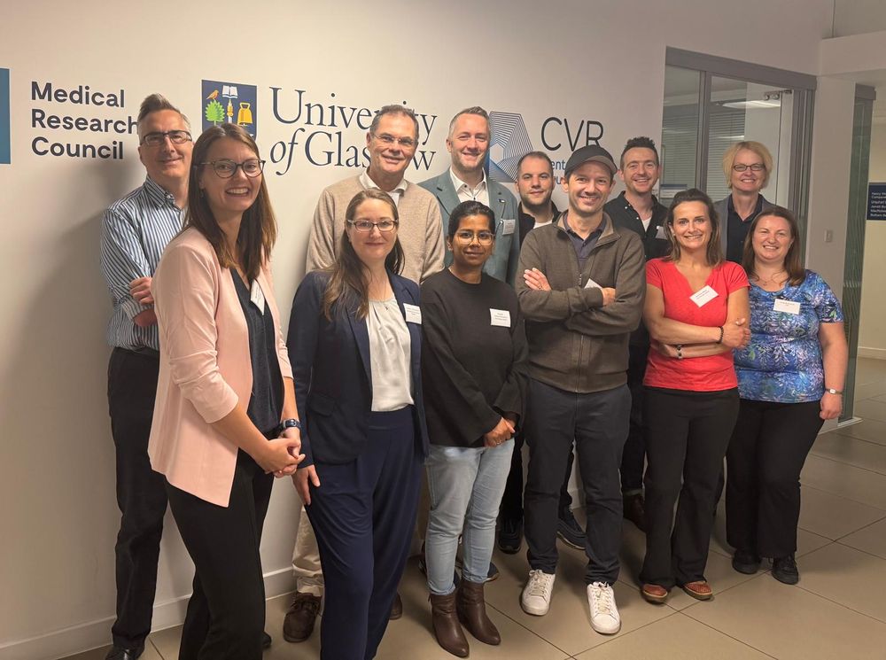 Twelve people pose for a group photo in front of a wall displaying logos for the Medical Research Council- University of Glasgow Centre for Virus Research. They are dressed in business casual attire. The setting is an indoor office.