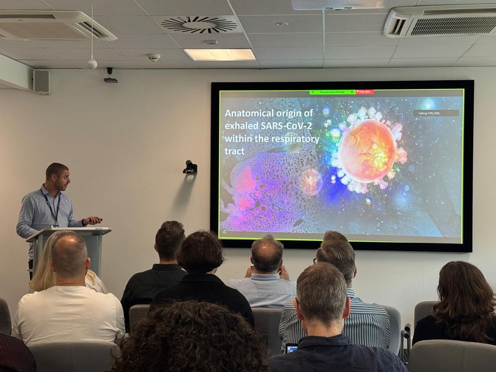  man presents to an audience in a conference room. The projected slide reads “Anatomical origin of exhaled SARS-CoV-2 within the respiratory tract” and features a colourful virus illustration. Attendees are seated and focused on the presentation.