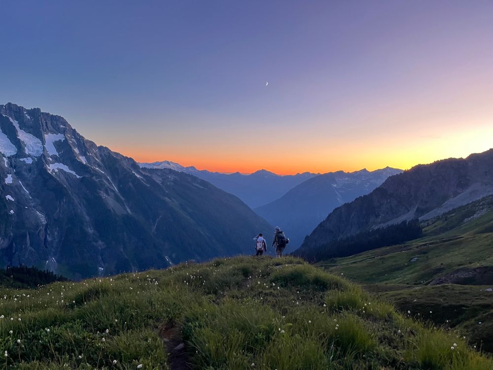 Two climbers descend lush green terrain at sunset, waking down a rise that is covered in lush grass. Their backs are to the camera. The imposing north face of a nearby peak rise with snow on its upper flanks. In the distance, the skyline of the Cascade mountains are a purple silhouette against the red, orange, and yellow sky. A crescent moon hangs above their head.