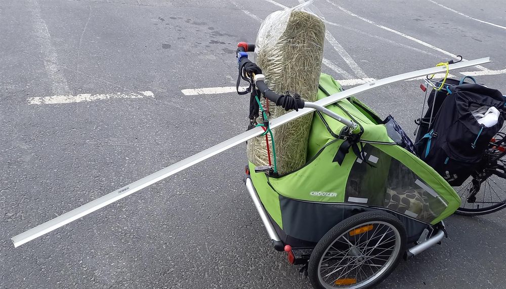 A bike trailer with a 1 metre sack of hay poking out of the top, and a 3 metre long piece of plastic protruding front and back.