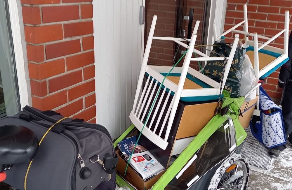 A bike trailer with kitchen chairs both the right way up and upside down, neatly stacked, extending about 50cm above and as much behind the dimensions of the trailer. Also bags and boxes in and hanging on the trailer.